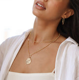 Woman wearing gold oval hoops and gold layering jewelry with a white background