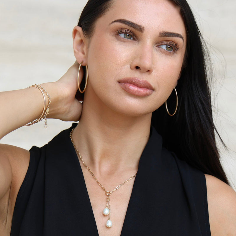 Woman wearing a black top and pearl necklace against a neutral background with Large Oval Gold Hoops
