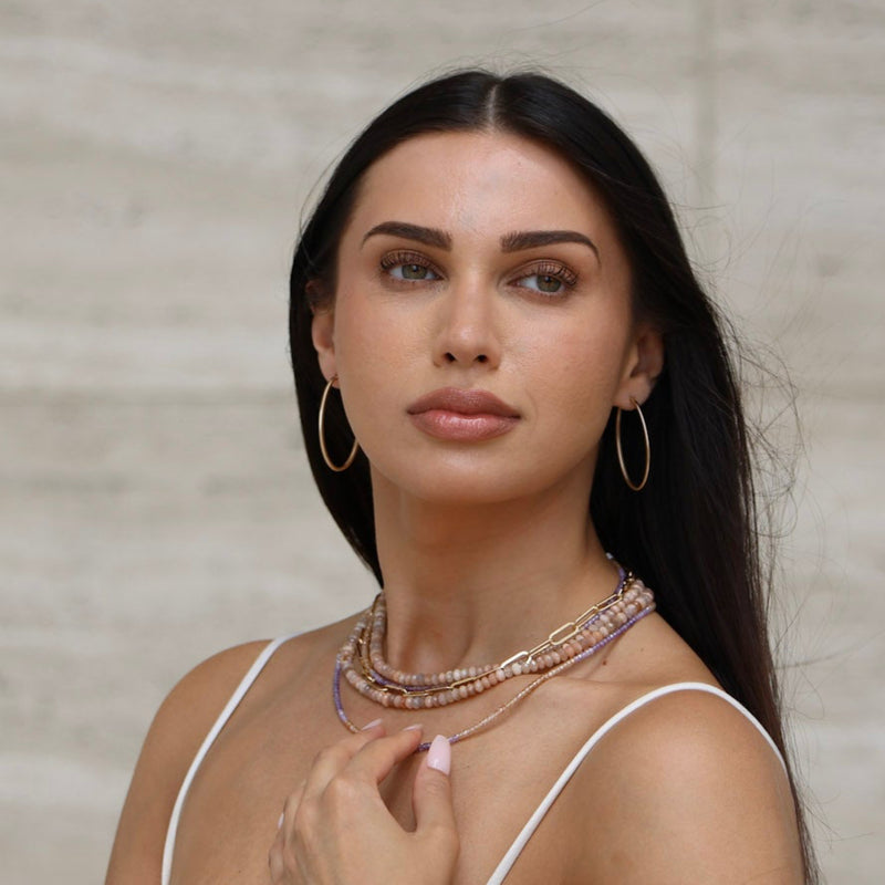 Woman wearing a white top and multiple necklaces against a neutral wall and large oval gold hoops