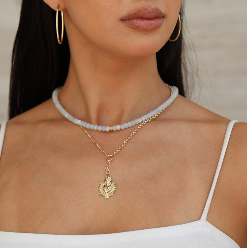 Woman wearing gold necklace with sacred heart  and hoop earrings against a neutral background
